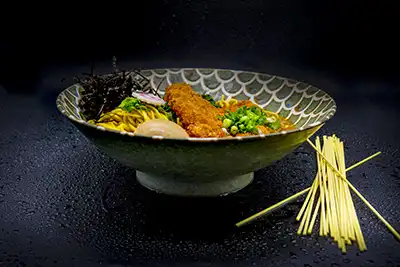 Caldo elaborado durante 24 horas a fuego lento con cerdo empanado Tonkatsu sobre caldo de ramen servido con verduras de temporada, tallarines, huevo, cebollino, Naruto laminado de pescado y algas.